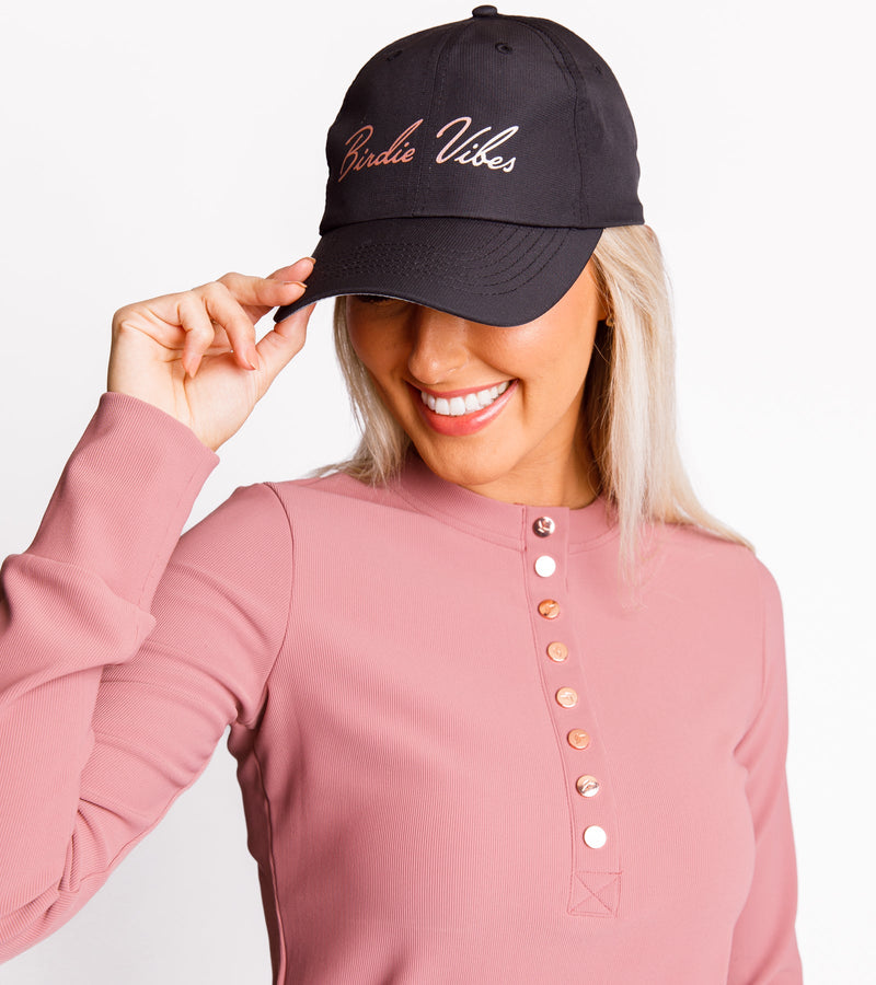 A woman wearing a black dad hat with rose gold foil printing that says "BIRDIE VIBES" and a pink long-sleeve shirt with rose gold buttons, adding a touch of elegance to her outfit.