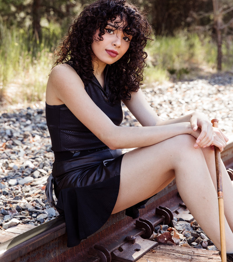 Sitting woman wearing a sleeveless black snakeskin polo with a stylish mock v-neck pop collar and a black skirt.