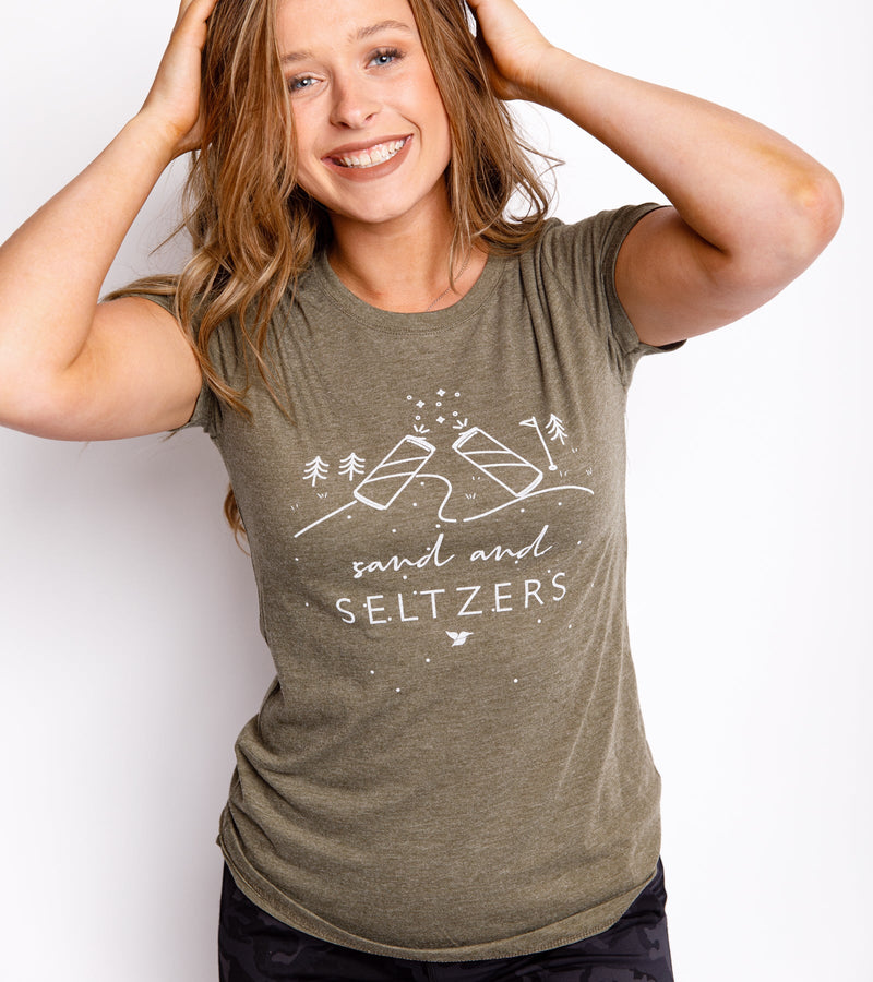 Smiling woman wearing an army green T-shirt with a white graphic that says "Sand and Seltzers."