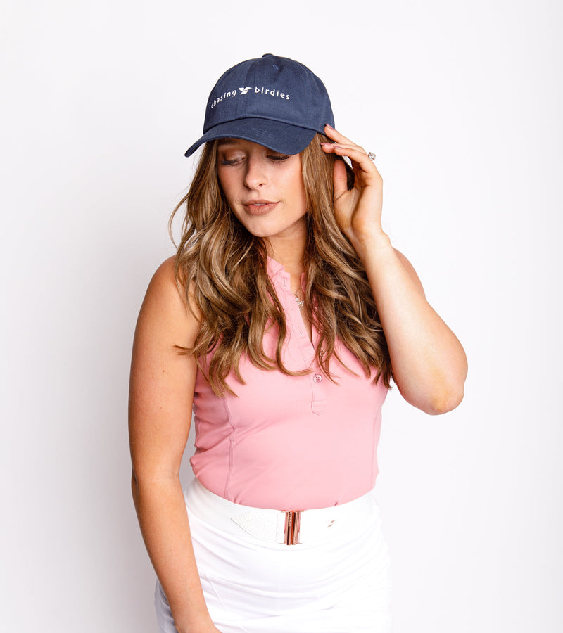  Woman wearing a navy dad hat with a white graphic saying "Chasing Birdies," a sleeveless pink top, and white bottoms.