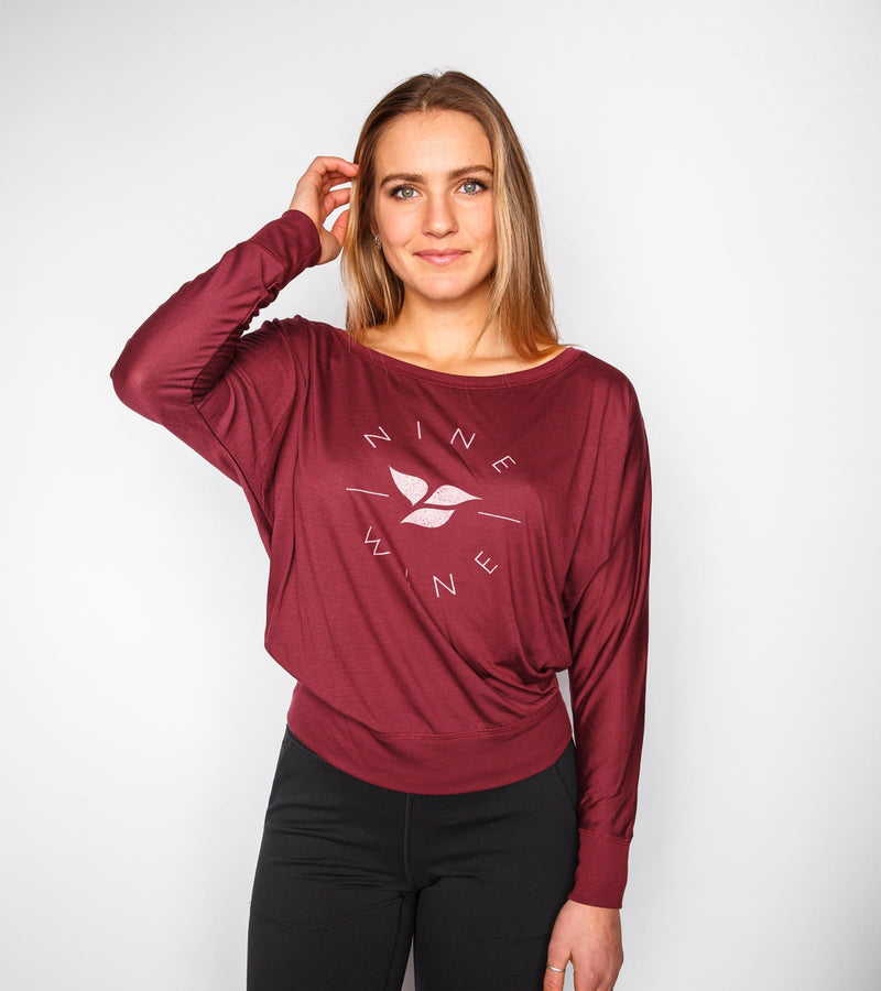 Woman wearing a long-sleeve maroon slouchy with pink graphics saying "NINE AND WINE."