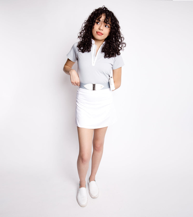 Woman wearing a short-sleeve gray polo with a front bonded zipper with white stitching and a placket. She also wears a gray and white belt with a white bag and a white skirt.