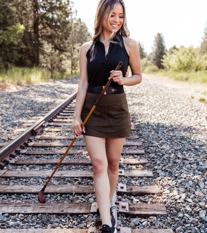 Front view of a woman wearing a sleeveless black polo with a deep front button placket, Princess stitch side seams for a more fitted look, a black belt, and an army green skirt while holding a golf club.