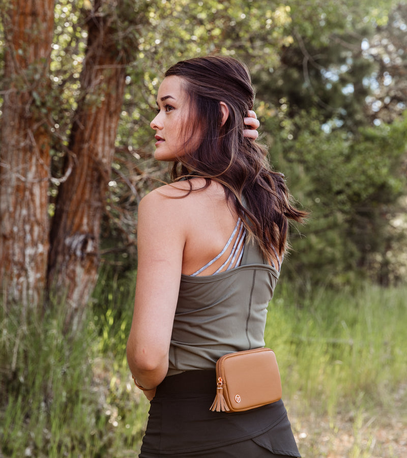  Back view of a woman wearing a sleeveless green army polo with a T-back design, a black belt with a honey purse, and black bottoms.