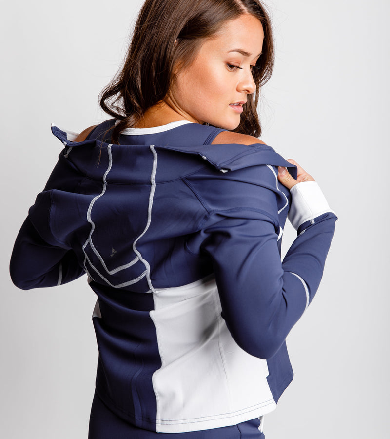 Close-up back view of a woman putting on a navy jacket with white sides and chevron stitching on the back.